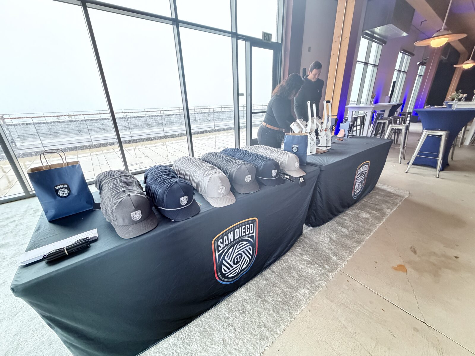 Patch bar table lined with trucker hats and compact presses at a glass-walled waterfront venue